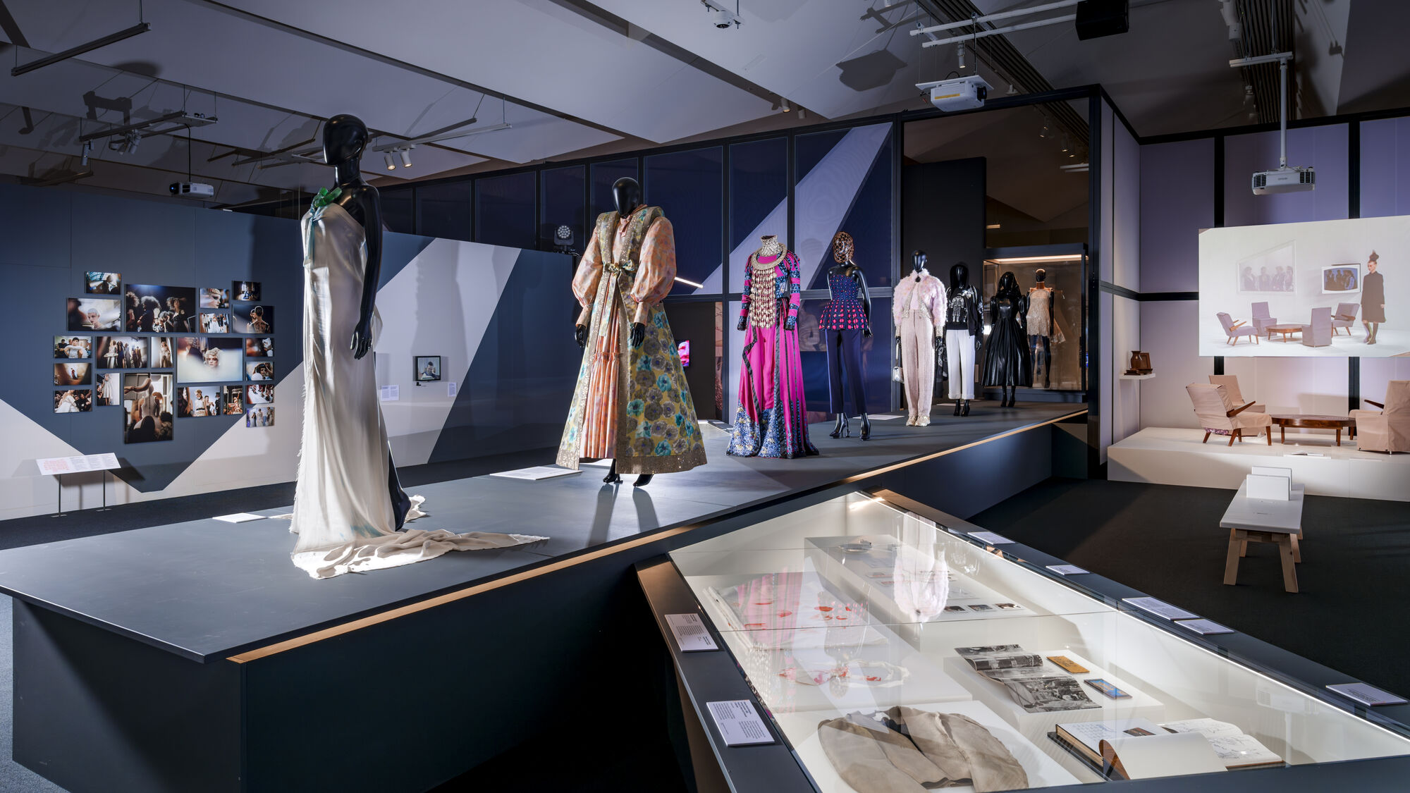 Fashion exhibition with mannequins in colourful garments by designers such as Maison Margiela and Viktor & Rolf, framed photos on the wall, and display cases with documents and artifacts.